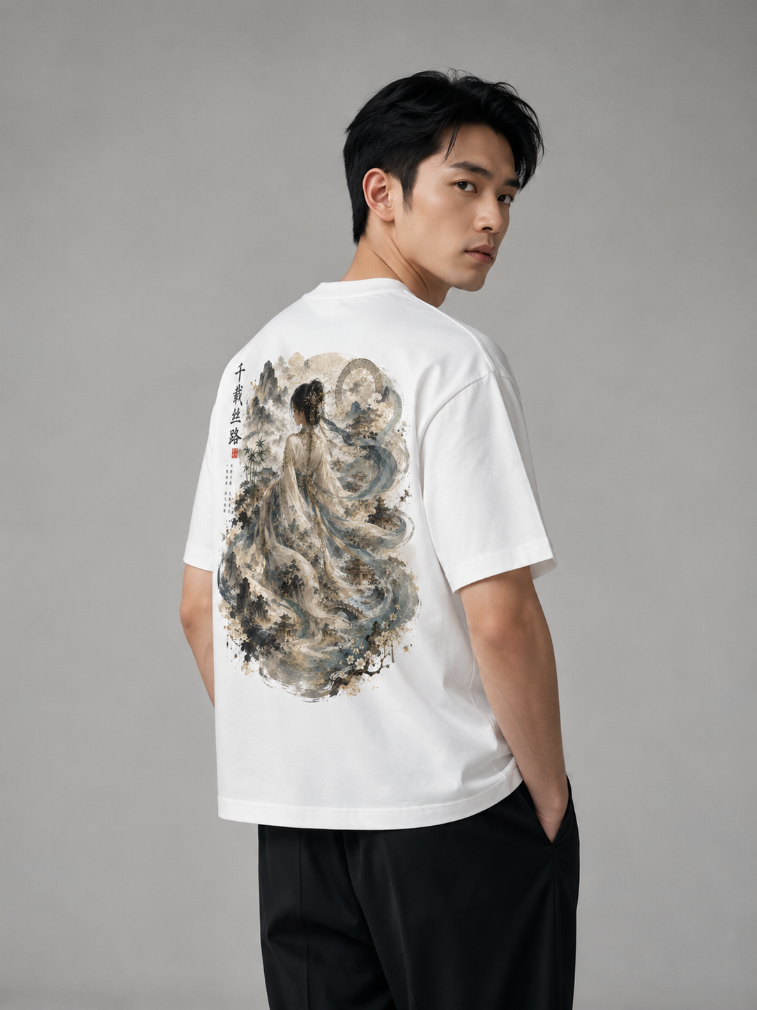A man wearing a white short-sleeve T-shirt with a large traditional Chinese ink-wash style print on the back, paired with black pants, photographed against a neutral gray studio background.