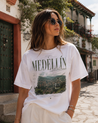 A woman wearing a white graphic t-shirt with 'MEDELLÍN' and a scenic print, paired with cream pants, standing outdoors on a cobblestone street in front of colonial-style buildings.
