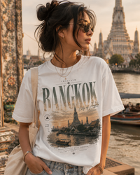 A woman wearing a white graphic t-shirt with a 'Bangkok City of Lights' print, standing outdoors near a river with a temple in the background.