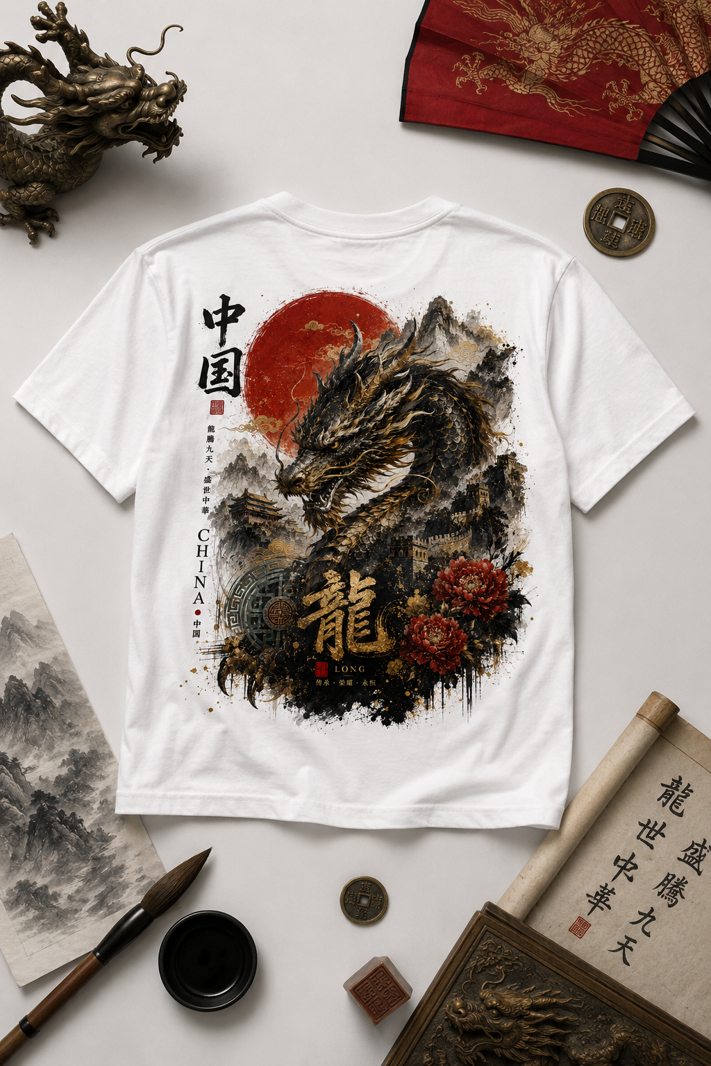 A white short-sleeve T-shirt laid flat on a light surface, featuring a large back graphic of a black dragon over a red sun and dark ink-style mountains, with Chinese characters and floral details. The shirt is styled with decorative props including a folding fan, coin, brush, ink stone, and rolled paper.