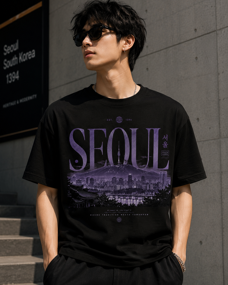 A man wearing a black graphic t-shirt with a purple Seoul cityscape print, standing outdoors against a gray concrete wall.