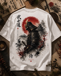 A white short-sleeve T-shirt laid flat on a styled surface, showing a large back graphic of a samurai warrior in black armor in front of a red sun and dark ink-style mountains. Japanese text appears on the shirt, and the scene includes decorative props like a folding fan, brush, and calligraphy paper around it.